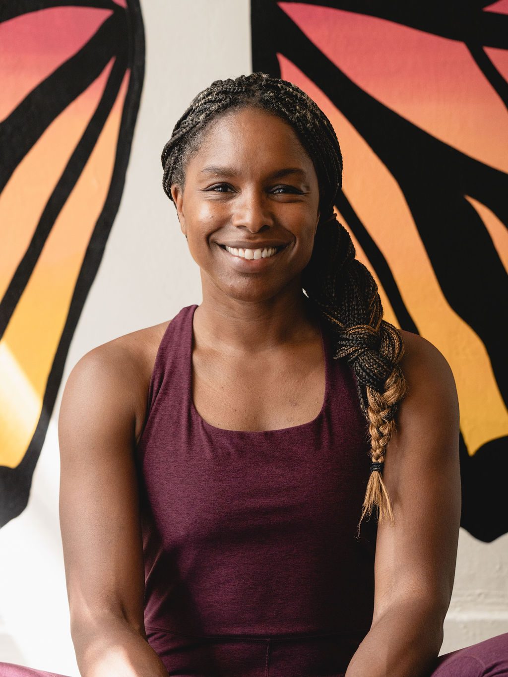 Tameka seated in front of the butterfly mural, smiling, braid over one shoulder.