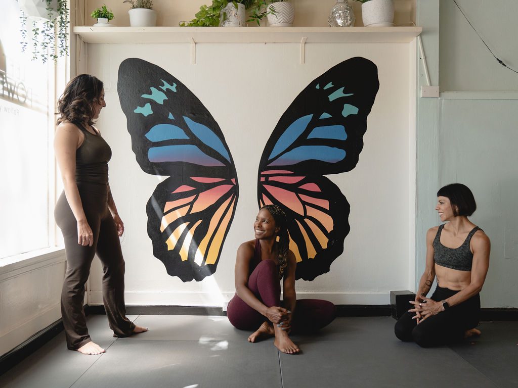 Three yogis laughing together on the mats beneath the full butterfly mural.