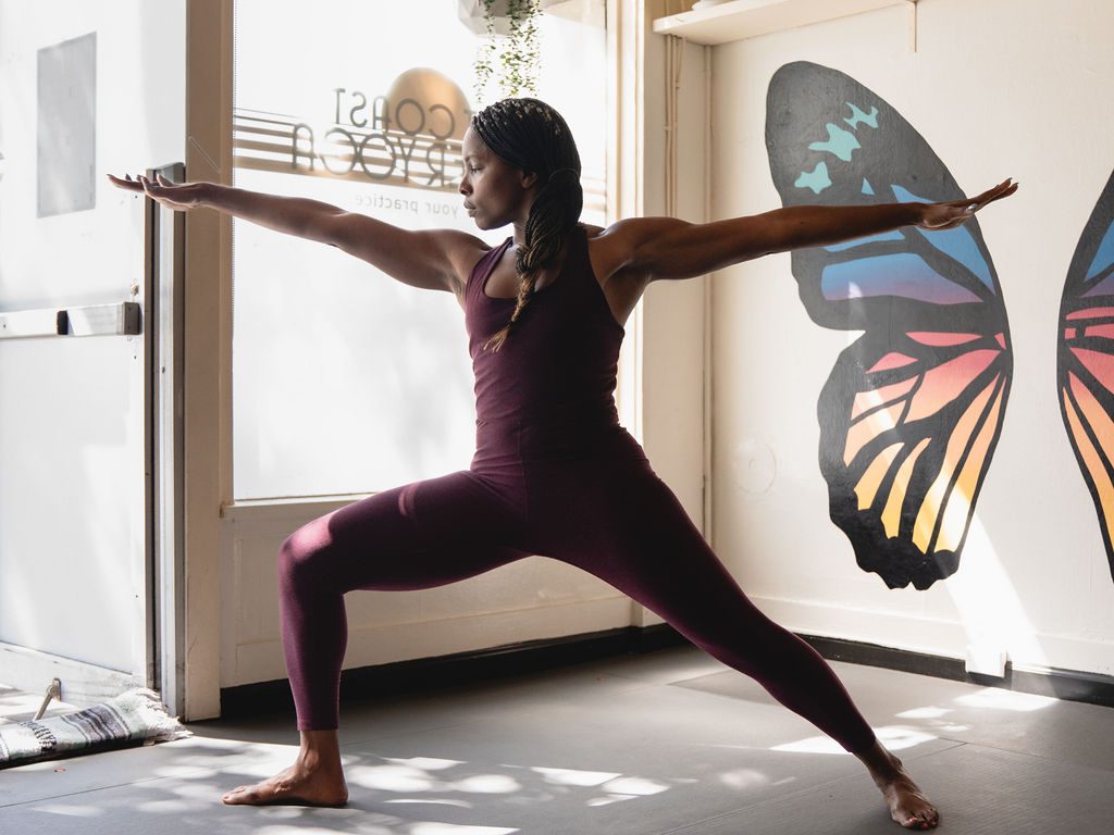 Tameka holds Warrior II in golden afternoon light at Left Coast Power Yoga, arms extended in front of a butterfly wing mural.