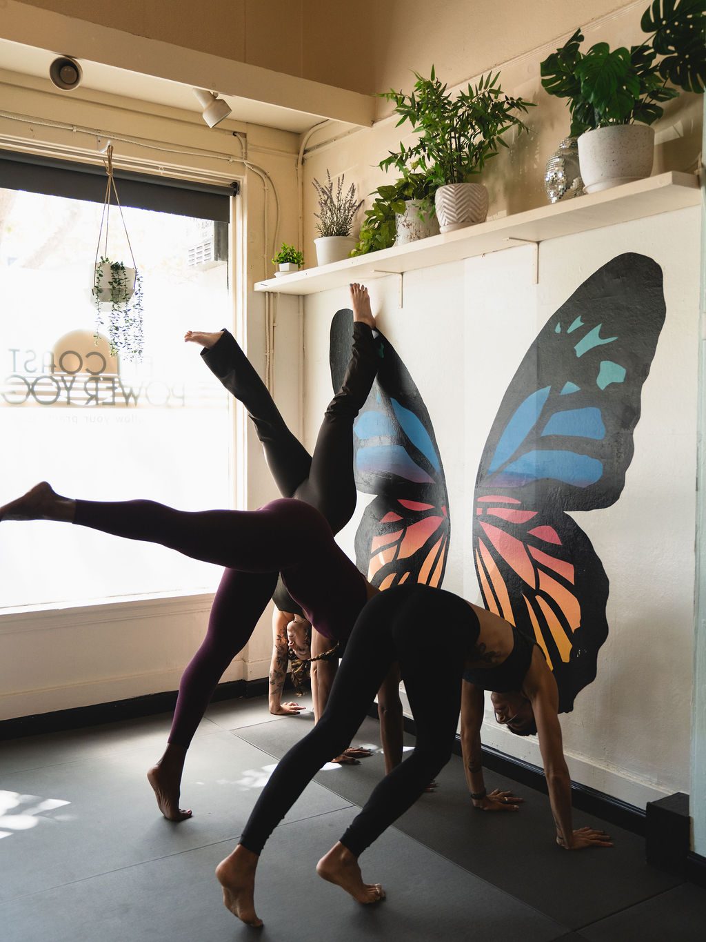 Three yogis in a row kicking up toward handstand at the wall, legs lifted, in front of the butterfly mural.